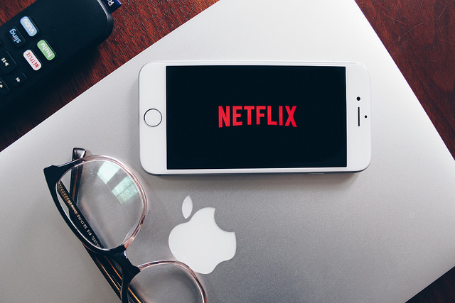 Image depicting a closed Apple laptop, with a pair of glasses and a smartphone resting on top. The phone is showing the Netflix logo.