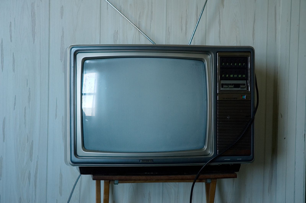 Image of an old fashioned television set. It is turned off, and light relfects against its blank grey screen.