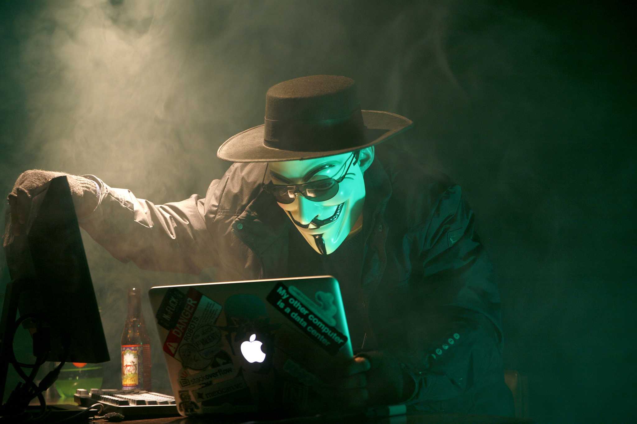 A person in a dark hat and trench coat peers down at two computer monitors. They are wearing a white mask, hiding their identity. The room is dark, and smoke floats in the air.