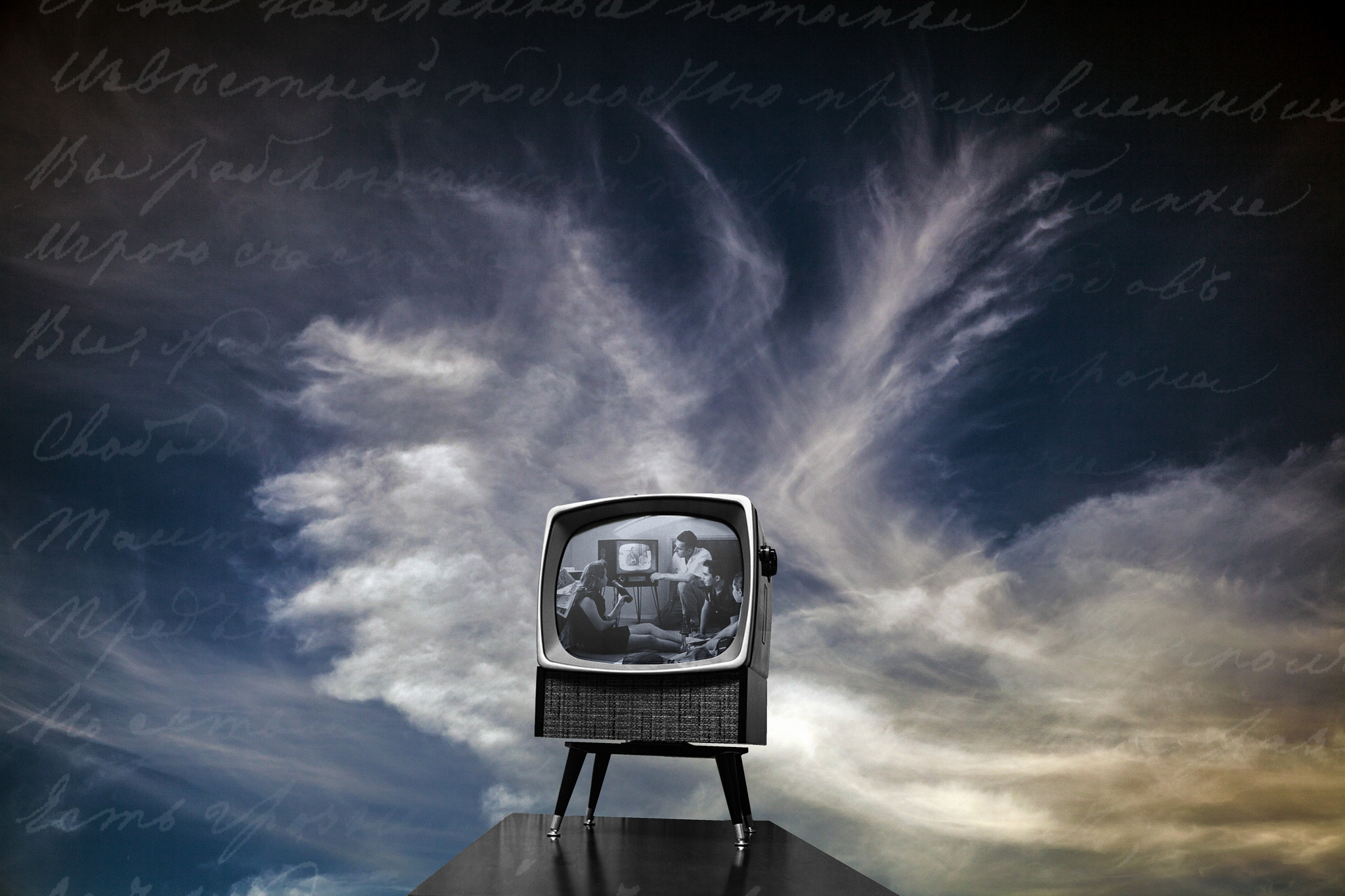 Photograph of an old-fashioned television set, up against the background of a cloudy sky. On the screen, a black and white image of a family watching television.