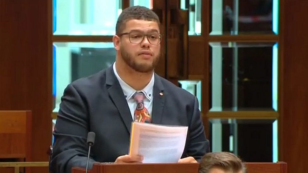 Senator Steele-John presents the bill in parliament. He looks ahead with a serious expression, holding the document in his hand. He wears a dark suit, glasses, and a tie depicting bright indigenous art. 