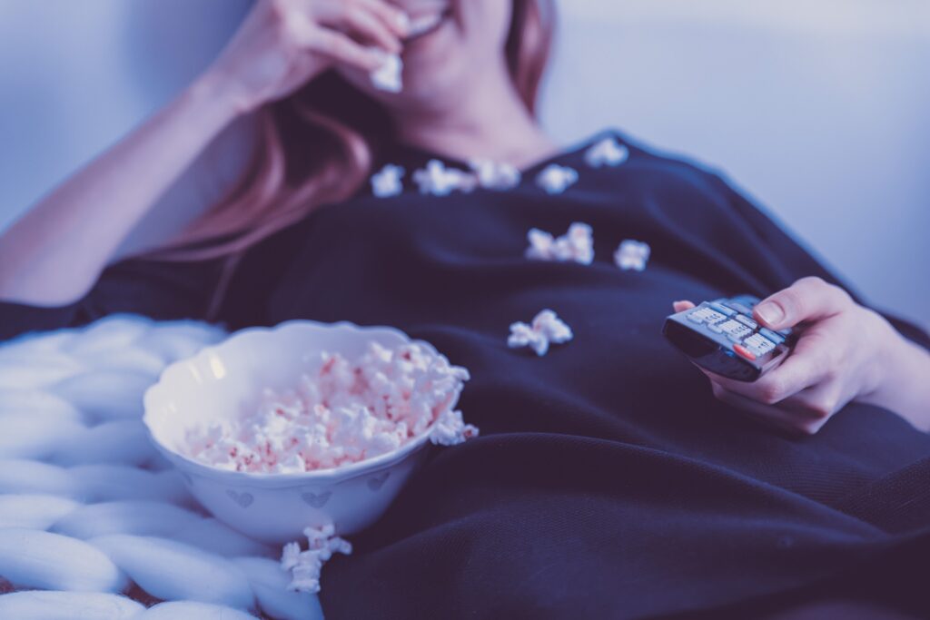 Image of a woman messily eating popcorn while watching television, holding a remote control in her hand.