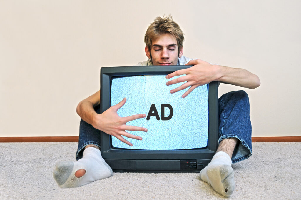 Photograph of a man hugging an old television set with the letters AD on the screen. His eyes are blissfully closed and his old socks have holes in them.